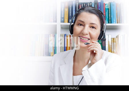 Freundlich lächelnde Frau mit Kopfhörern und weißen Blazer vor Bücher, in einer Videokonferenz, Customer Support Agent oder helpline Assistant Stockfoto