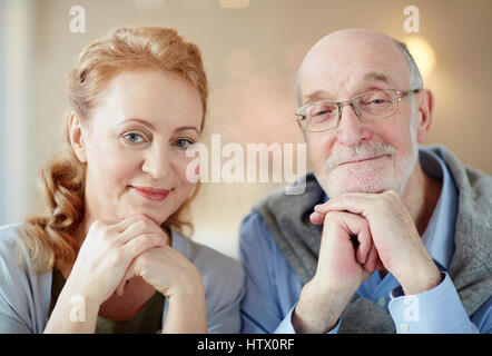 Porträt von nett lächelnd älteres paar zu Hause, graue dunkelhaarigen Mann und elegante ältere Frau Blick in die Kamera ruht Kopf auf Händen Stockfoto