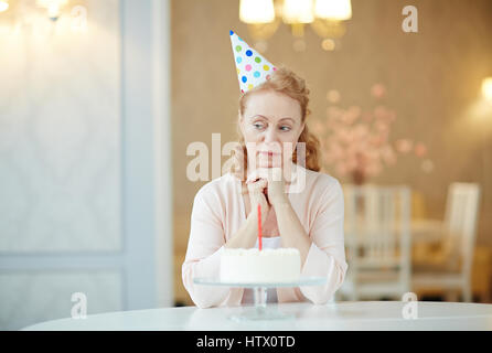 Porträt von melancholischen Reife Frau sitzt alleine am Geburtstagstisch mit Kuchen, tragen Partyhut und suchen traurig Stockfoto