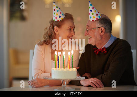 Porträt von senior Liebespaar feiert Geburtstag zusammen sitzen am Tisch mit Kuchen und tragen Partyhüte suchen Sie zärtlich in die Augen Stockfoto