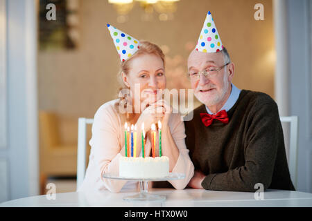 Porträt von senior Liebespaar feiert Geburtstag zusammen sitzen am Tisch mit Kuchen und tragen Partyhüte lächelnd und Blick in die Kamera Stockfoto