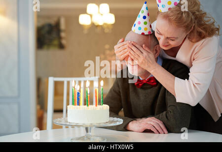 Porträt von senior Liebespaar Geburtstag feiern, Reife Frau, die geschlossenen Augen lächelnder alten Mann präsentiert ihn Geburtstagstorte mit Kerzen Stockfoto