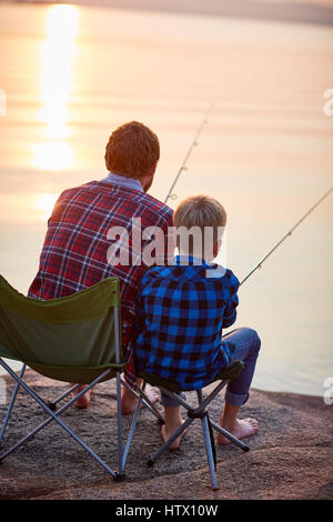 Rückansicht-Porträt von Vater und Sohn sitzen zusammen auf Felsen mit Stangen in ruhiger See Fischen Gewässer im Abendlicht, beide tragen karierten Hemden Stockfoto