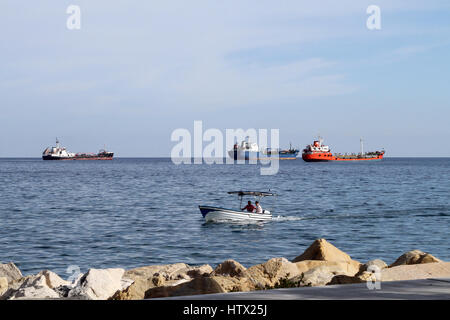 Ein kleines Boot hereinkommt zum Ufer entlang der Küste von Limassol, Zypern, mit großen Tankern und anderen Schiffen im Hintergrund. Stockfoto