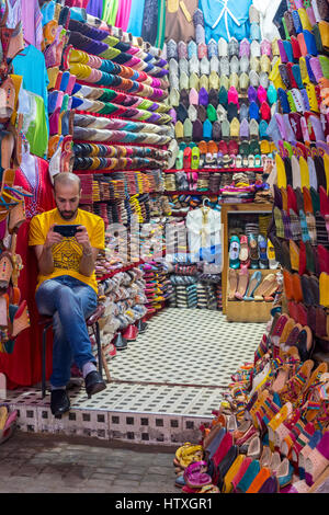 Fes, Marokko.  Hersteller von Schuhen und Sandalen Überprüfung sein Handy.  Tala'a Kabira Straßenmarkt, Fes El-Bali. Stockfoto