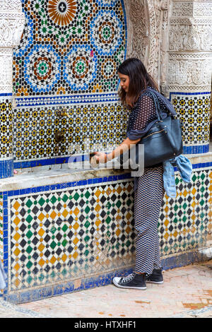 Fes, Marokko.  Nejjarine zu platzieren.  Junge Frau mit öffentlichen Trinkbecher am Brunnen. Stockfoto