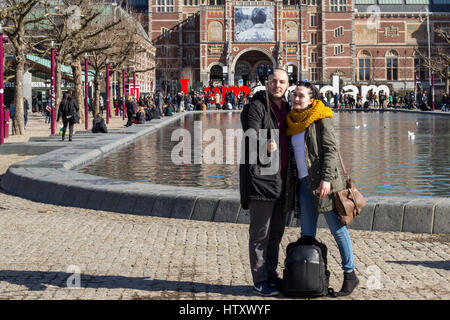 Junge Touristen-paar unter ein Selbstporträt in Amsterdam mit einem iPhone auf ein Selfiestick mit dem Amsterdam-Zeichen und das Rijksmuseum auf dem Hintergrund Stockfoto