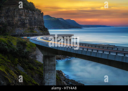 Sonnenuntergang über der Sea Cliff Bridge entlang der australischen Pazifikküste Ozean mit Lichtern der vorbeifahrenden Autos in der Nähe von Sydney, Australien. Langzeitbelichtung. Stockfoto