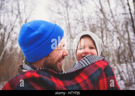 Porträt eines Vaters, der sein Kind in einem Winterwald in einer Decke gehüllt trägt Stockfoto