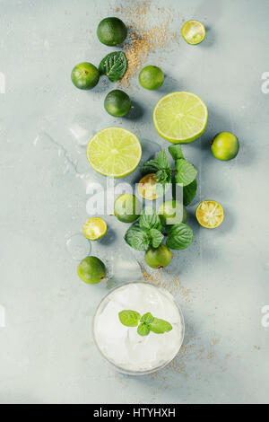 Zutaten für Mojito cocktail, ganze, in Scheiben geschnittenen Limetten und Mini-Limetten, Minze, brauner Kristallzucker über graue Steinstruktur Hintergrund mit Glas ful Stockfoto