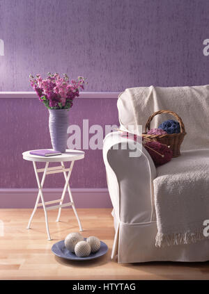 Vase mit rosa Lager auf einem kleinen weißen Tisch neben einem weißen Sofa mit einem Korb aus Wolle und Stricknadeln in einem lila Wirtschaft-Wohnzimmer Stockfoto
