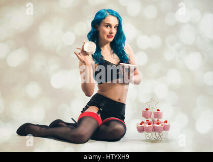 Freche Pin-up Mädchen trägt Nylons und Strapsen halten ein Vintage Tasse und Untertasse mit Kirsch Cupcakes an ihrer Seite Stockfoto