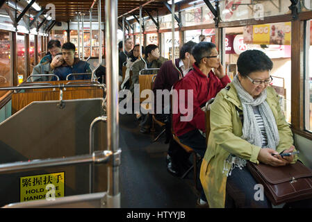 Passagiere sitzen in einer Straßenbahn Fahrt entlang einer Straße in Causeway Bay mit einer Frau, die ihr Handy überprüfen. Stockfoto