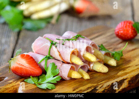 Rollen von gekochten Schinken mit frischen weißen Spargel gebunden mit Schnittlauch und serviert auf einem Holzbrett mit Erdbeeren Stockfoto