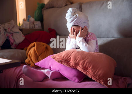 Kleines schüchternes Mädchen mit Handtuch auf Kopf auf Sofa mit Kissen sitzen und Gesicht mit Händen versteckt. Stockfoto