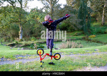 6-jähriger Junge Kind Kind Gleichgewicht auf einem Bein auf umgedrehtem Fahrrad und machen taub Geste spielen in Country Garden Wales UK KATHY DEWITT Stockfoto