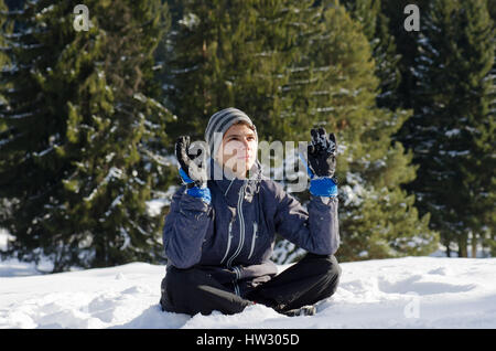 Jugendlicher Mann sitzen auf Schnee mit Winterjacke und Handschuhe betrachten und denken in Yogaposition mit Tannen hinter Stockfoto