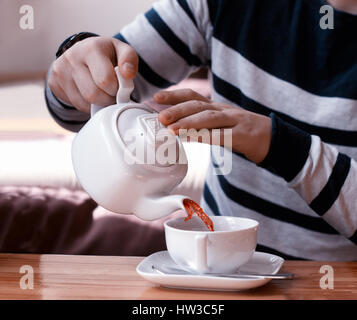 Man gießt Früchtetees von Teekanne in die weiße Tasse auf Holztisch Stockfoto