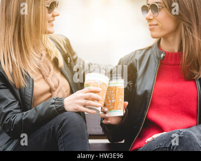 Porträt von zwei schönen jungen Freundinnen sitzen auf Bank und Pappbecher Kaffee in Händen halten. Stockfoto