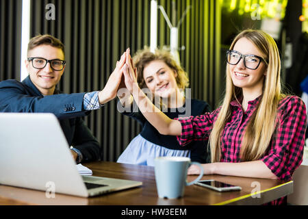 Drei Studenten jubeln, geben hohe fünf miteinander glücklich nach erfolgreichen Präsentation ihres gemeinsamen Projekts an der Universität, im Café sitzen wi Stockfoto