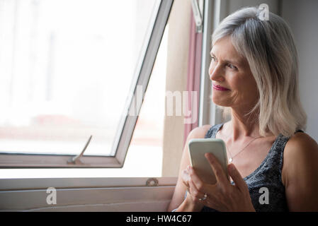 Lächelnde kaukasischen Frau SMS auf Handy in der Nähe von Fenster Stockfoto