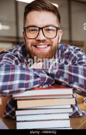 Lächelnde männlichen Studenten ruht auf Stapel Bücher beim Sitzen am Schreibtisch in Bibliothek Stockfoto