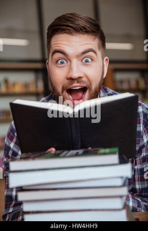Porträt von aufgeregt männlichen Studenten Lesebuch hinter Stapel Bücher auf Bibliotheksschalter Stockfoto