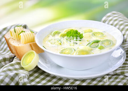 Cremige vegetarische Käse und Lauch Suppe mit gebuttertem Baguette, Lauch im Hintergrund Stockfoto