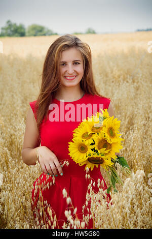 Porträt von schönen Brünette Mädchen mit langen Haaren tragen im roten Kleid stehen unter Pikelets im Feld. Junge weibliche Holding Blumenstrauß von denselben lächelnd Stockfoto