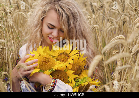 Hübsche junge Ukrainerin mit lockigem Haar in ethnischen Nationaltrikot riechen Blumenstrauß von Sonnenblumen und Lächeln auf den Lippen tragen. Schöne blonde weibliche stehend Stockfoto