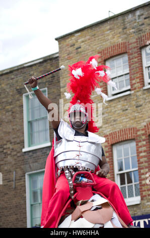 Karneval in Hackney ein schwarzer Mann hält ein Stöckchen wie ein römischer Soldat in einem Kostüm Weste und einem flammenden rot Kopfschmuck Stockfoto