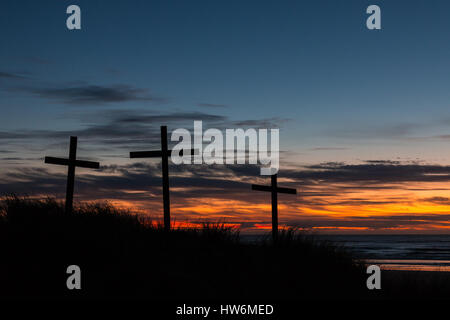 Drei Kreuze auf einer Sanddüne mit einem Sonnenuntergang über dem Meer. Stockfoto