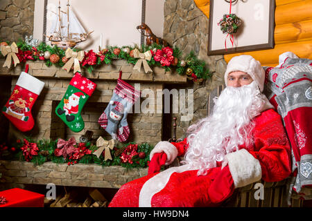 Bärtigen Weihnachtsmann in einem Sessel sitzend, Geschenk-Boxen, Kamin und dekorierten Weihnachtsbaum im Hintergrund Stockfoto