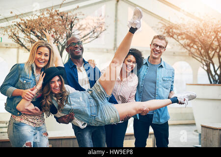 Gruppe von Jugendlichen, die Hände vor ihnen ihr Freund Mädchen festhalten, posieren, Kamera und lachen, drinnen im warmen Licht stehen Stockfoto