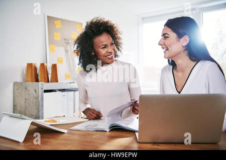 Zwei glückliche attraktive junge Multirassischen Unternehmerinnen gemeinsam lachen als sie sitzen in einem hellen high key Office mit Papierkram und einem Laptop compu Stockfoto