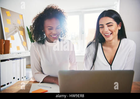 Zwei motivierte junge Unternehmerinnen lachend, als sie auf einem Laptop-Computer auf ihren neuen kleinen Startup-Unternehmen zusammenarbeiten Stockfoto