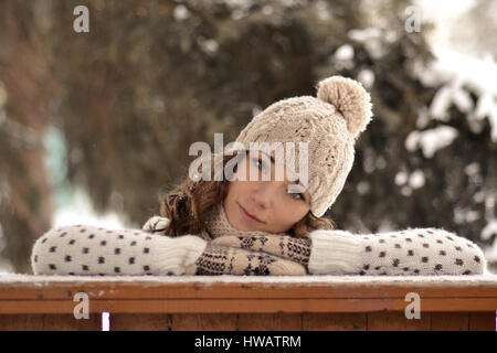 Sehr schöne, schöne Mädchen mit Hut, Pullover, warme Handschuhe, lockigen, braunen Haare im Winter verschneiten Wald aussehen, Sie, kuscheln, Kopf auf die Hände legen, setzen Stockfoto