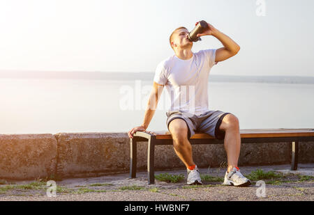 junge starke kaukasischen Mann mit weißen Kopfhörer Getränke einen Protein-Cocktail aus einem Shaker nach dem Ausführen unter freiem Himmel. Die helle Sonne leuchtet es. Stockfoto