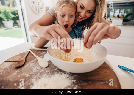Mutter und Tochter machen Teig in Küche. Frau, die den Teig und kleine Mädchen mit Staunen Ausdruck Ei hinzufügen. Stockfoto