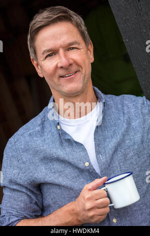 Porträt gedreht von einem attraktiven, erfolgreichen und glücklichen Mann mittleren Alters männlichen trägt ein blaues Hemd trinken Tee oder Kaffee aus einer Blechtasse Stockfoto
