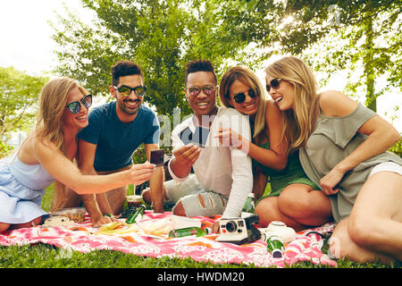 Fünf Erwachsene Freunde betrachten Film aus Sofortbild-Kamera beim Picknick im park Stockfoto