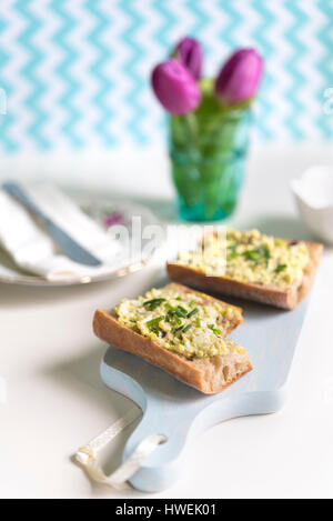Nahaufnahme von frischem Baguette-Sandwich mit Eiersalat auf blauen Schneidebrett mit Haufen von Tulpen im Hintergrund mit verteilt. Stockfoto