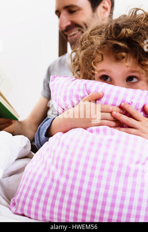 Mädchen Kissen umarmt, während Vater Märchenbuch im Bett liest Stockfoto