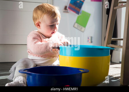 Babymädchen sitzen auf Küchenboden spielt mit Schalen Stockfoto