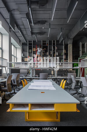 Lange grau-gelb Tisch mit gelben Stühle im Büro im Loftstil. Es gibt graue Arbeitsplätze mit Computern, Regale mit Büchern und roten Leiter, Stockfoto