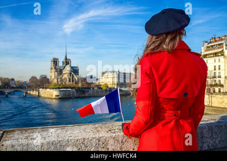 Hell in Paris. Hinter modernen Reisenden Frau im roten Mantel am Ufer in der Nähe von Notre Dame de Paris in Paris, Frankreich mit französischen Flagge gesehen Stockfoto
