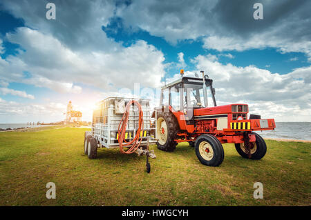 Roter Traktor mit Anhänger auf der Wiese gegen Leuchtturm und blau bewölktem Himmel im Frühjahr bei Sonnenuntergang. Landwirtschaftliche Zugmaschine. Landwirtschaftliche Maschinen ein Stockfoto