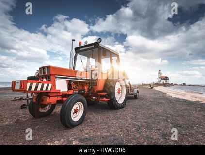 Roter Traktor auf der Wiese gegen Leuchtturm und blau bewölktem Himmel im Frühjahr bei Sonnenuntergang. Landwirtschaftliche Zugmaschine. Landwirtschaftliche Maschinen und Maschinen ich Stockfoto