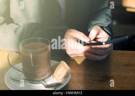 Frau mit Handy und trinken heißen Schokolade im Café. Lässige jungen kaukasischen Erwachsenfrau hinter dem Glas Textmitteilung zu schreiben, auf Smartphone ap Stockfoto