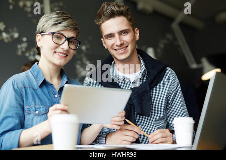 Niedrigen Winkel Ansicht von zwei Kollegen sitzen in kleinen gemütlichen Café und Blick in die Kamera mit einem breiten Lächeln, Pappbecher Kaffee und Laptop auf Tisch stehend Stockfoto